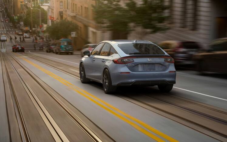 Rear driver-side view of the 2023 Honda Civic Sport Hatchback in Sonic Gray Pearl, driving on a city street.
