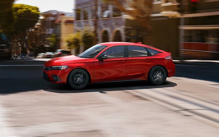 Driver-side profile view of the 2023 Honda Civic Sport Hatchback in Rallye Red, driving on a city street.