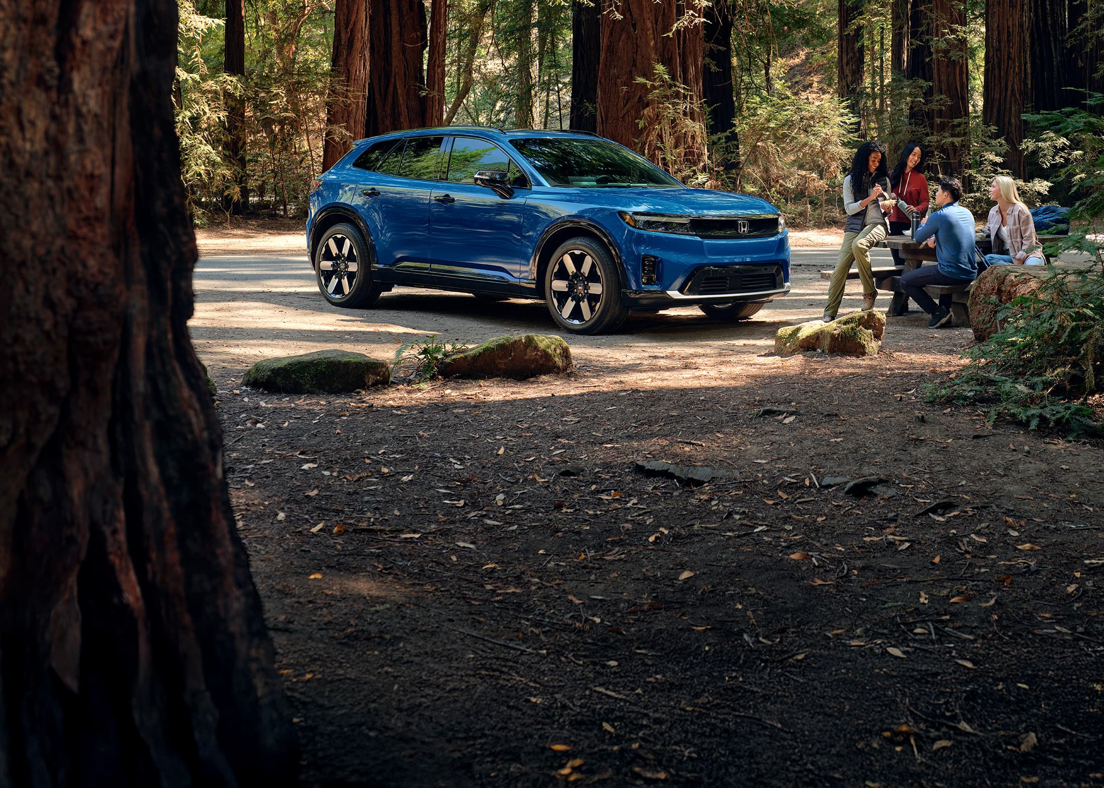 Front passenger view of the 2024 Honda Prologue Elite in Pacific Blue Metallic, shown parked with redwood trees behind it